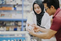 Asian woman in hijab shopping at a pharmacy, focused on finding products for her loved one’s needs.