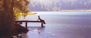 A person seated on a dock, gazing thoughtfully over a serene lake under a clear sky.