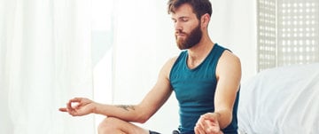 A man sits in a yoga pose on a bed, meditating peacefully, representing strength and mindfulness in difficult moments.