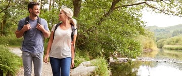 Two people hiking by a tranquil river with lush greenery surrounding them.