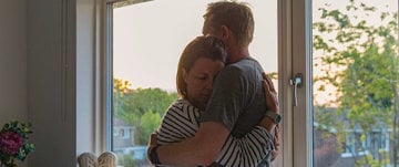 A couple shares a warm hug in front of a window, symbolizing love and intimacy in a serene setting.