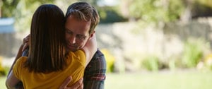 Two people hugging, one in a yellow top, in front of a garden.