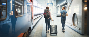 Two travelers with luggage walking towards a train platform, ready for their journey.