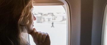 A woman gazes out an airplane window, contemplating her journey while managing migraine discomfort.