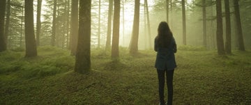 A woman stands amidst a lush forest, embodying resilience despite her ongoing battle with migraines since 2013.