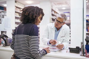 Inside a pharmacy, a man and woman discuss items while standing near shelves filled with various pharmaceutical products.