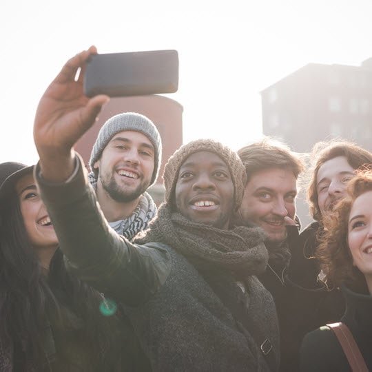 A diverse group of friends smiling and posing together for a selfie in a lively outdoor setting.