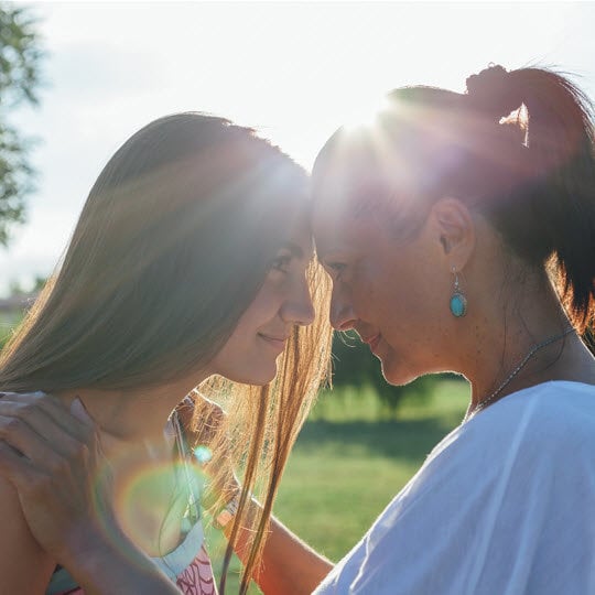 Two women share a warm embrace under the sunlight, radiating joy and friendship in a serene outdoor setting.