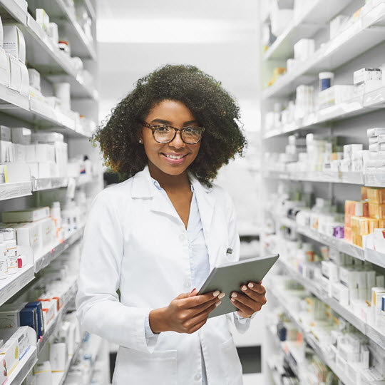 A woman in a white lab coat stands in a pharmacy aisle, representing professionalism in pharmacy practice and education.