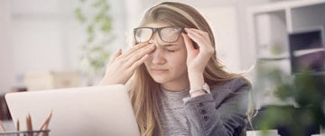 A woman with glasses sits at her desk, resting her head in her hands, reflecting the struggle of working with chronic migraine.