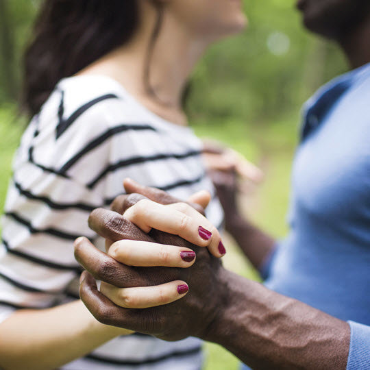 A man and woman hold hands while walking together in a serene park setting, surrounded by greenery.