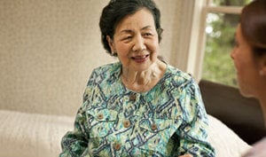 An elderly woman sits comfortably on a couch, accompanied by a caring nurse, highlighting health and wellness support.